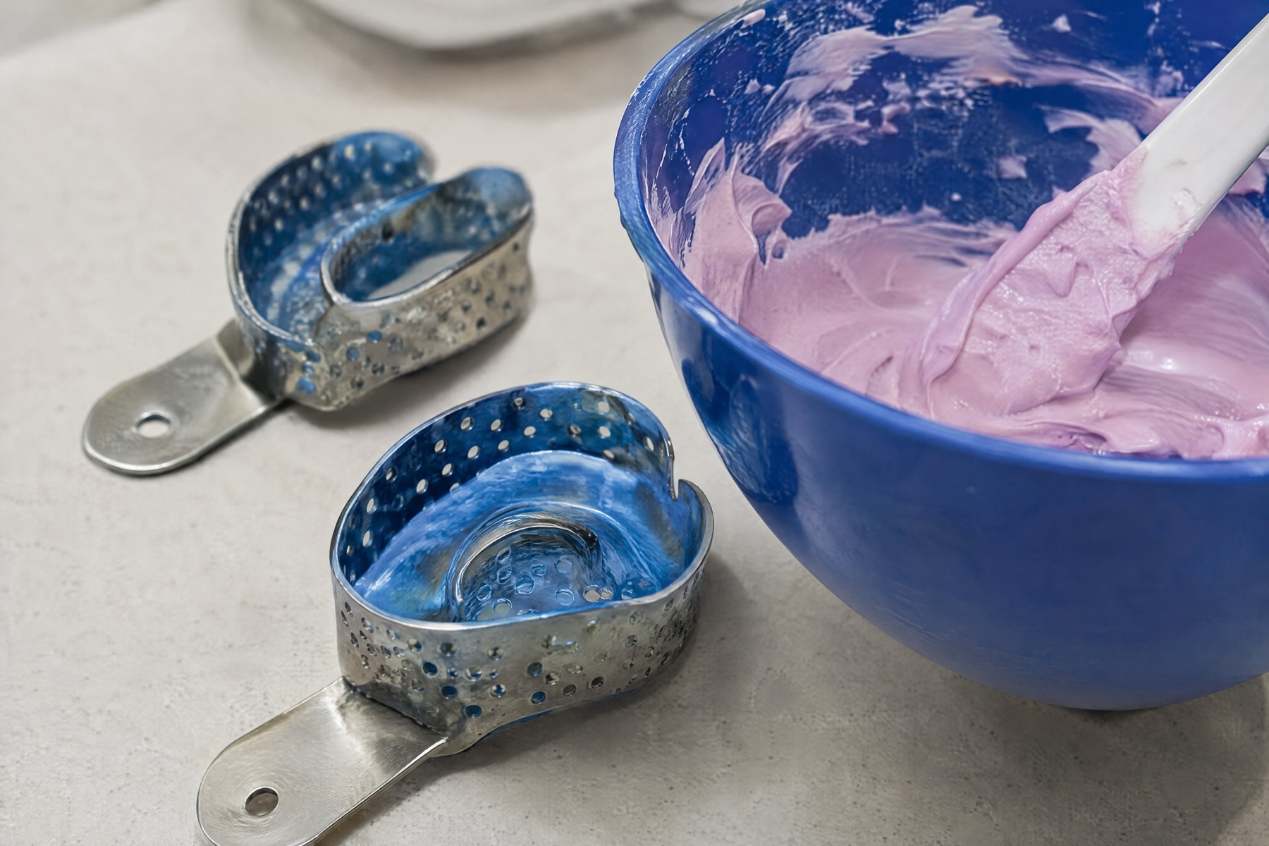Two perforated metal impression trays beside a blue mixing bowl of pink impression material